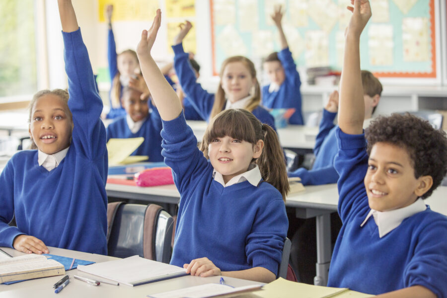 Pupils in a classroom at their desks. They all have their hands up.