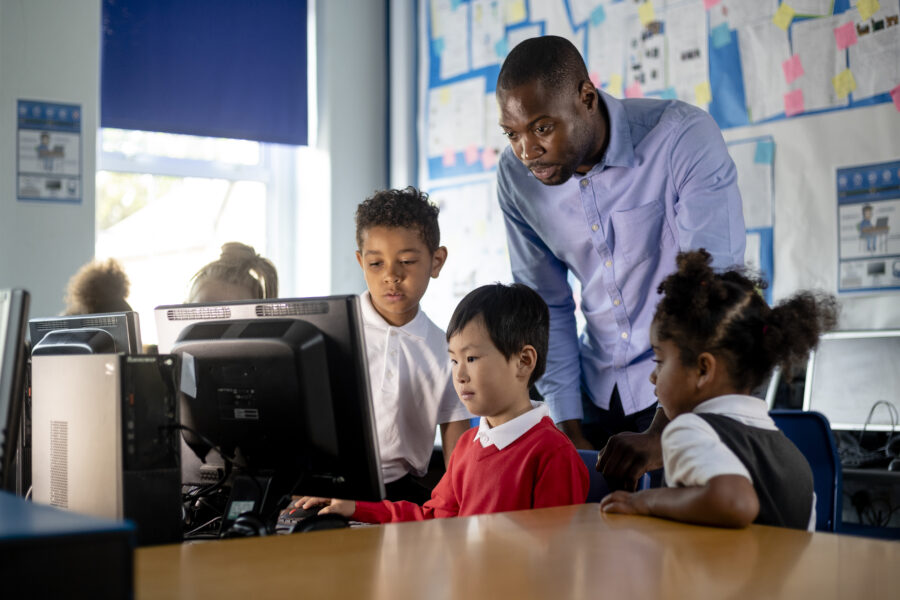 Three pupils in a classroom looking at a computer monitor with a teacher leaning over them